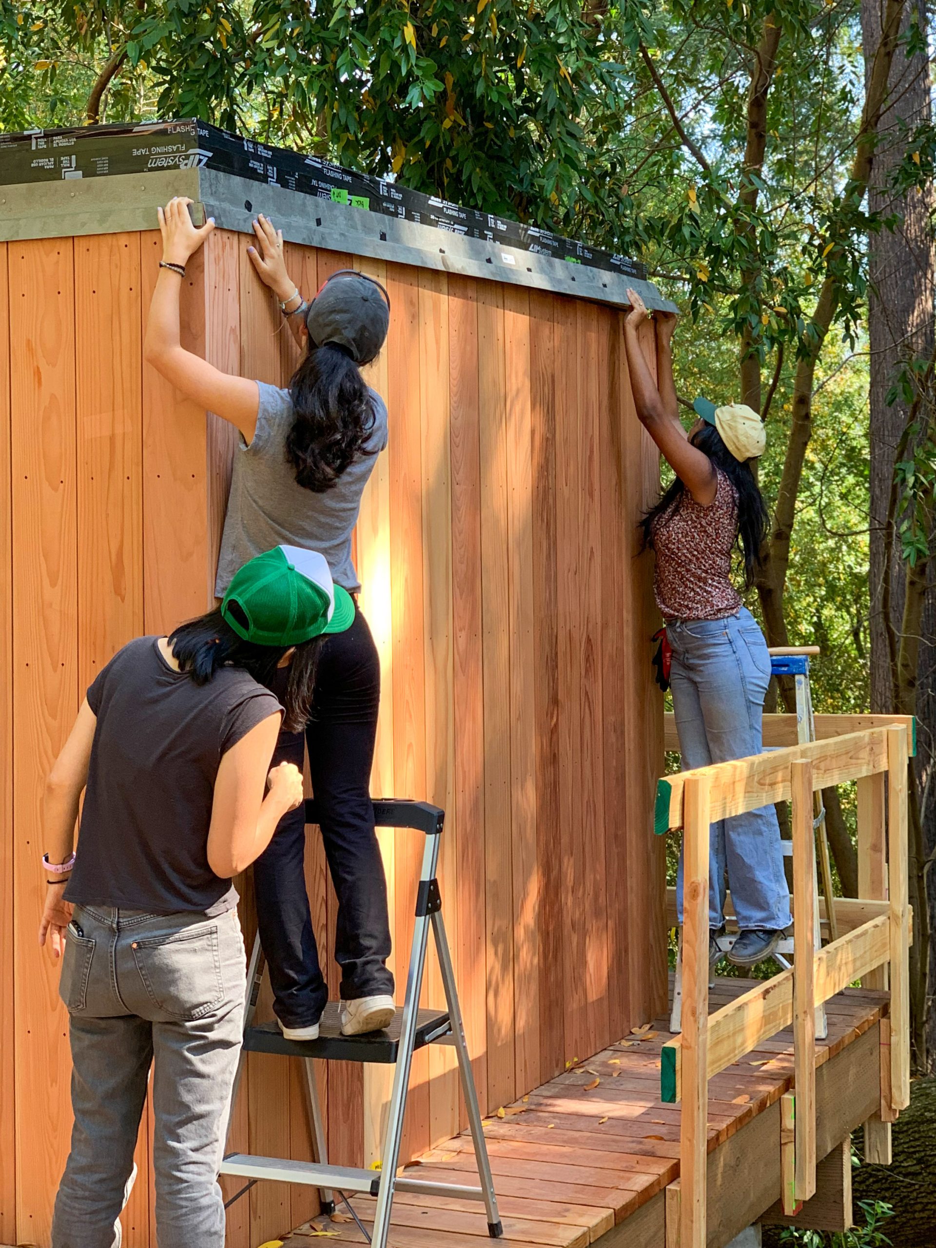 Three young women reaching up to apply flashing to a wooden outdoor sauna