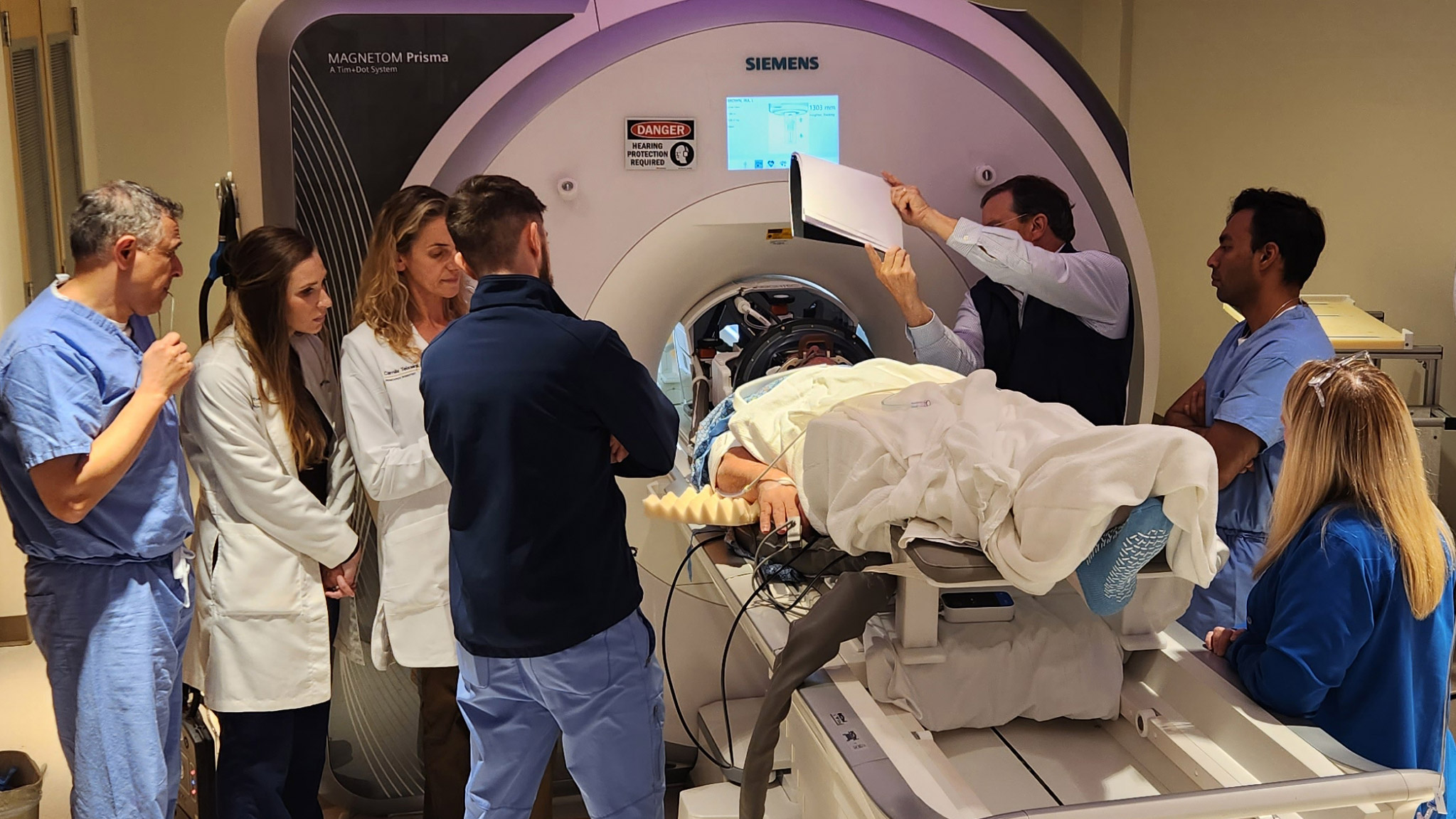 A patient surrounded by a medical team lays on the bed of an MRI machine with their head in a special focused ultrasound helmet